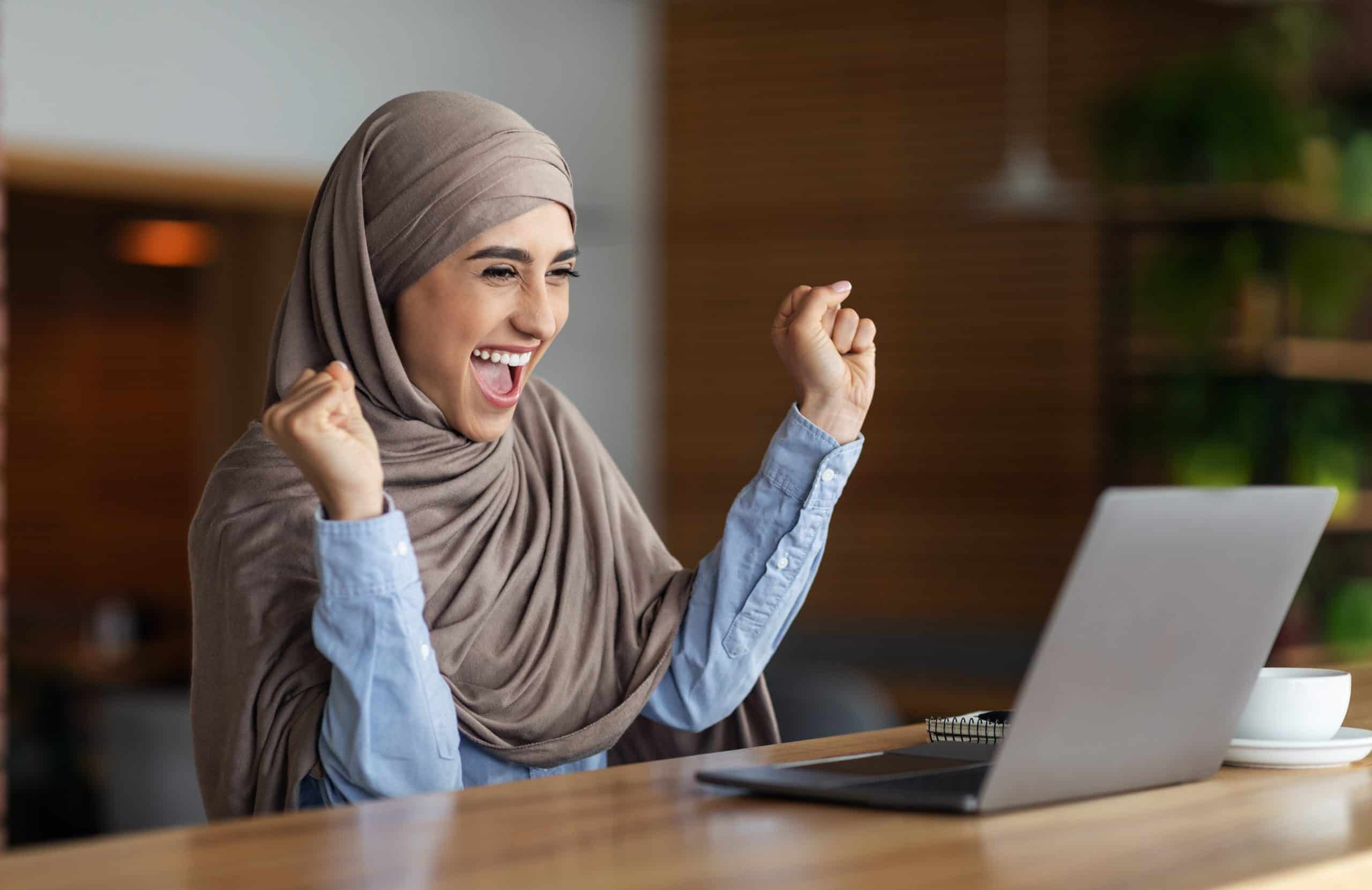 Emotional muslim woman in headscarf looking at laptop screen and raising hands up. Happy arab lady using laptop at cafe got good news, found nice job , empty space, career and job seeking concept